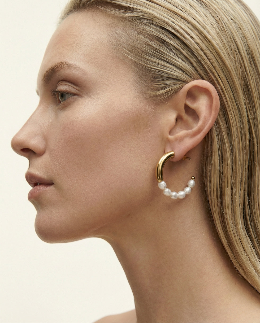 Close-up of a woman wearing gold hoop earrings with pearl-like beads on a neutral background