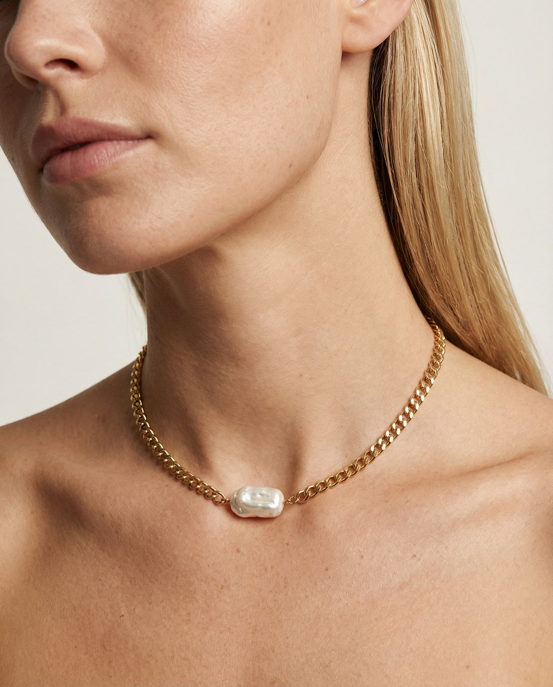 Close-up of a woman wearing a gold chain necklace with a pearl pendant on a neutral background
